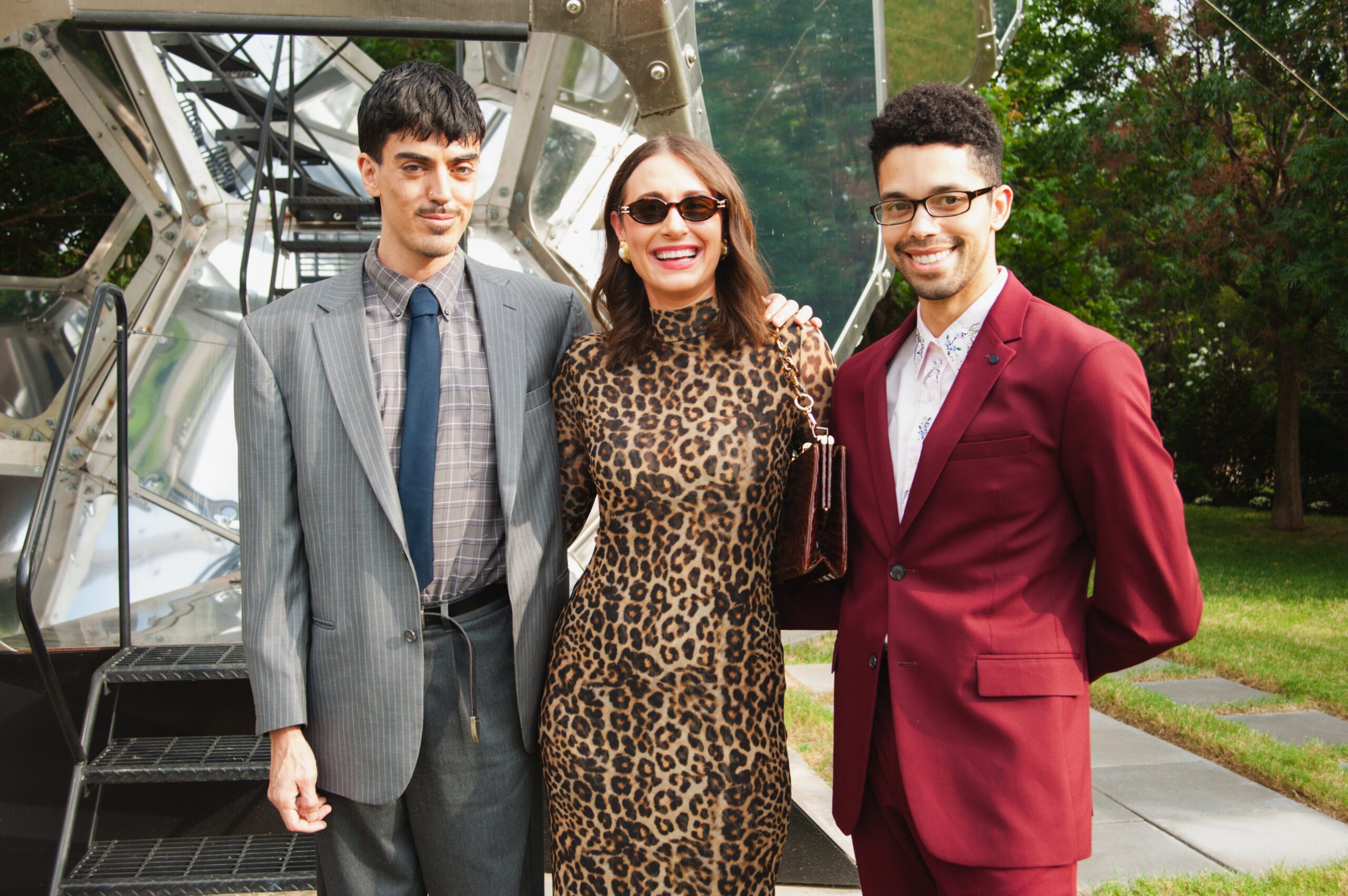 Two people in suits and a person in a leopard print dress post in front of a metal sculpture