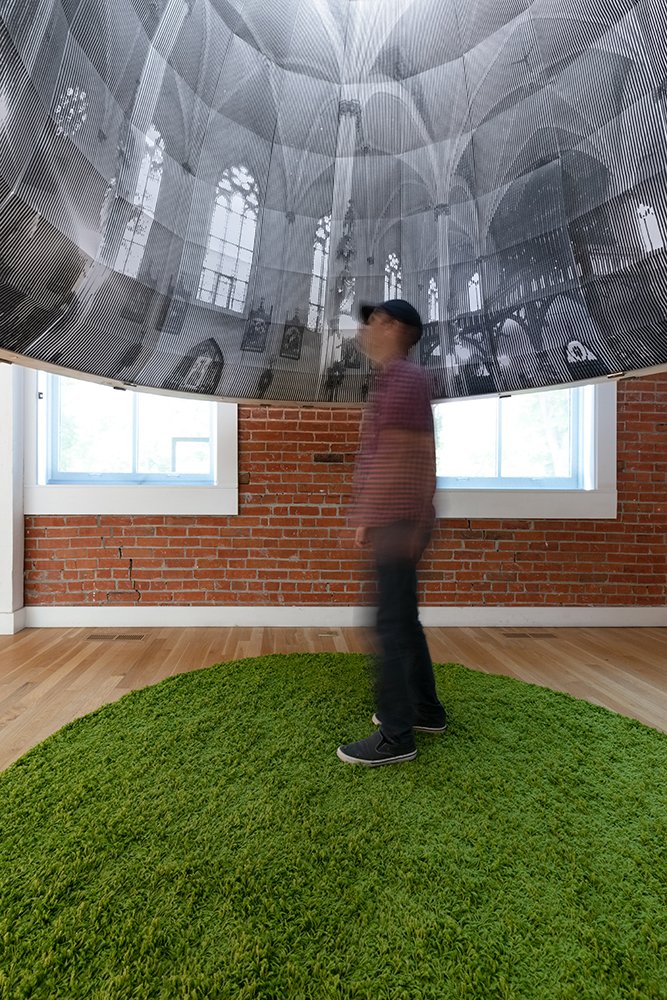 A man in a ballcap stands under a dome printed with an image of the inside of a church