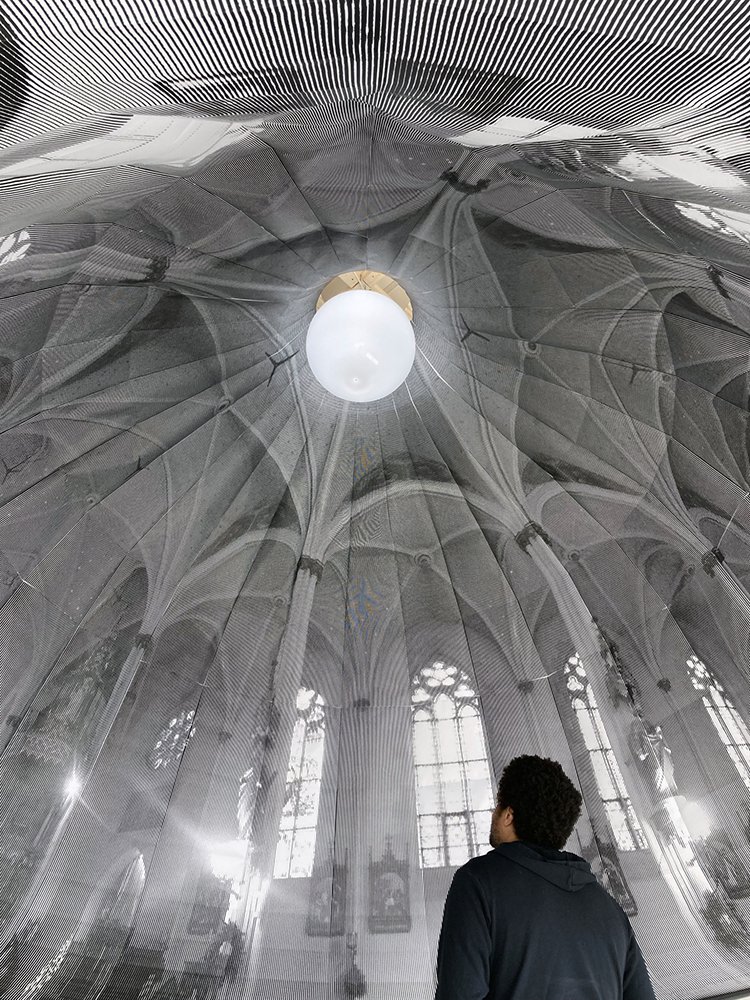 A Black man stand under a dome printed with an image of the inside of a church