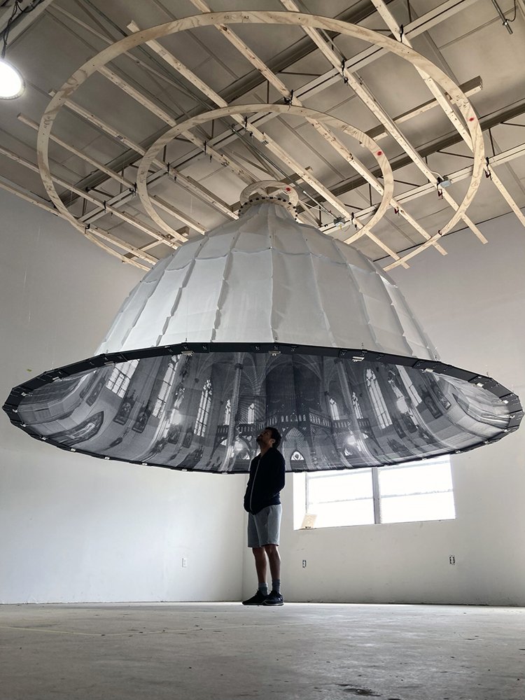 A Black man stands under a paper dome printed with the image of an inside of a church