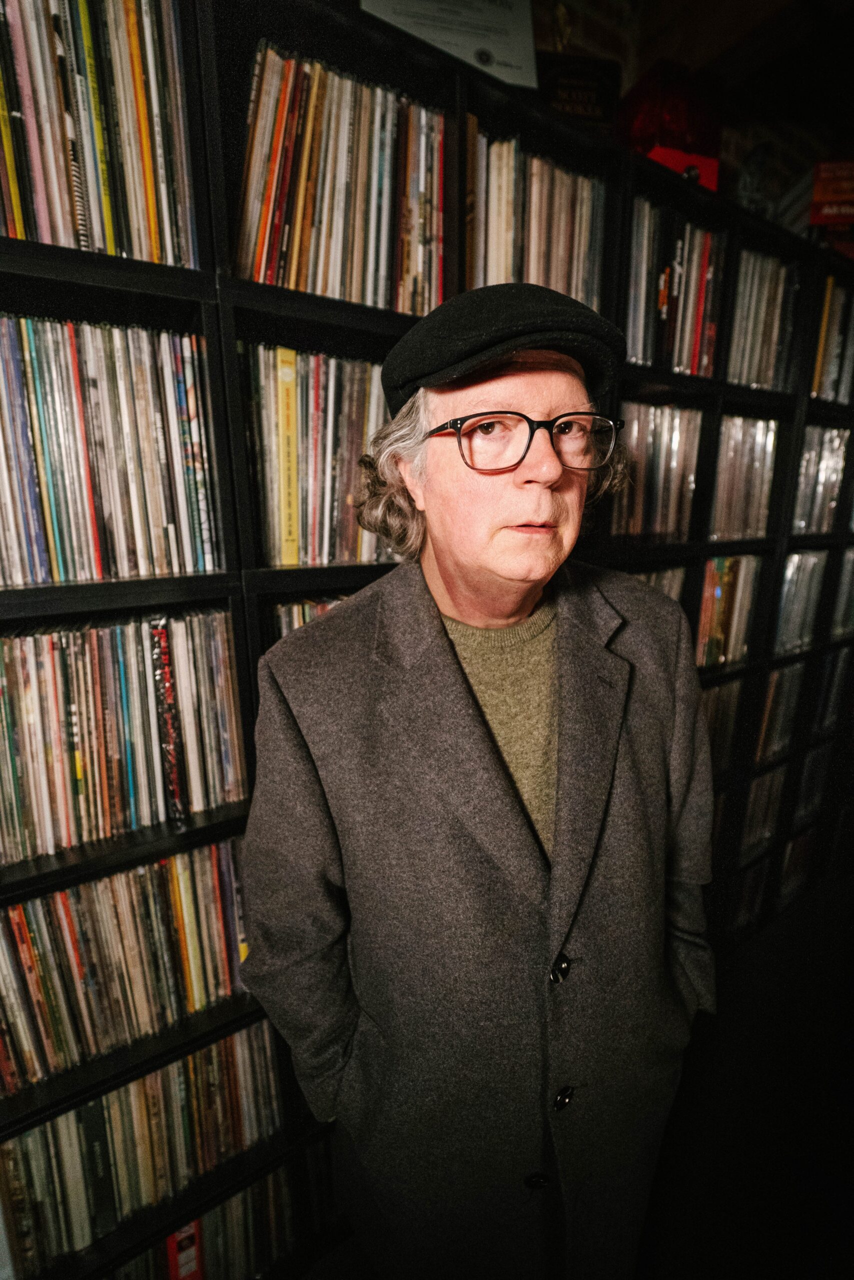 Scott Booker, a man wearing a flat cap and tweed coat, stands in front of a shelf of records