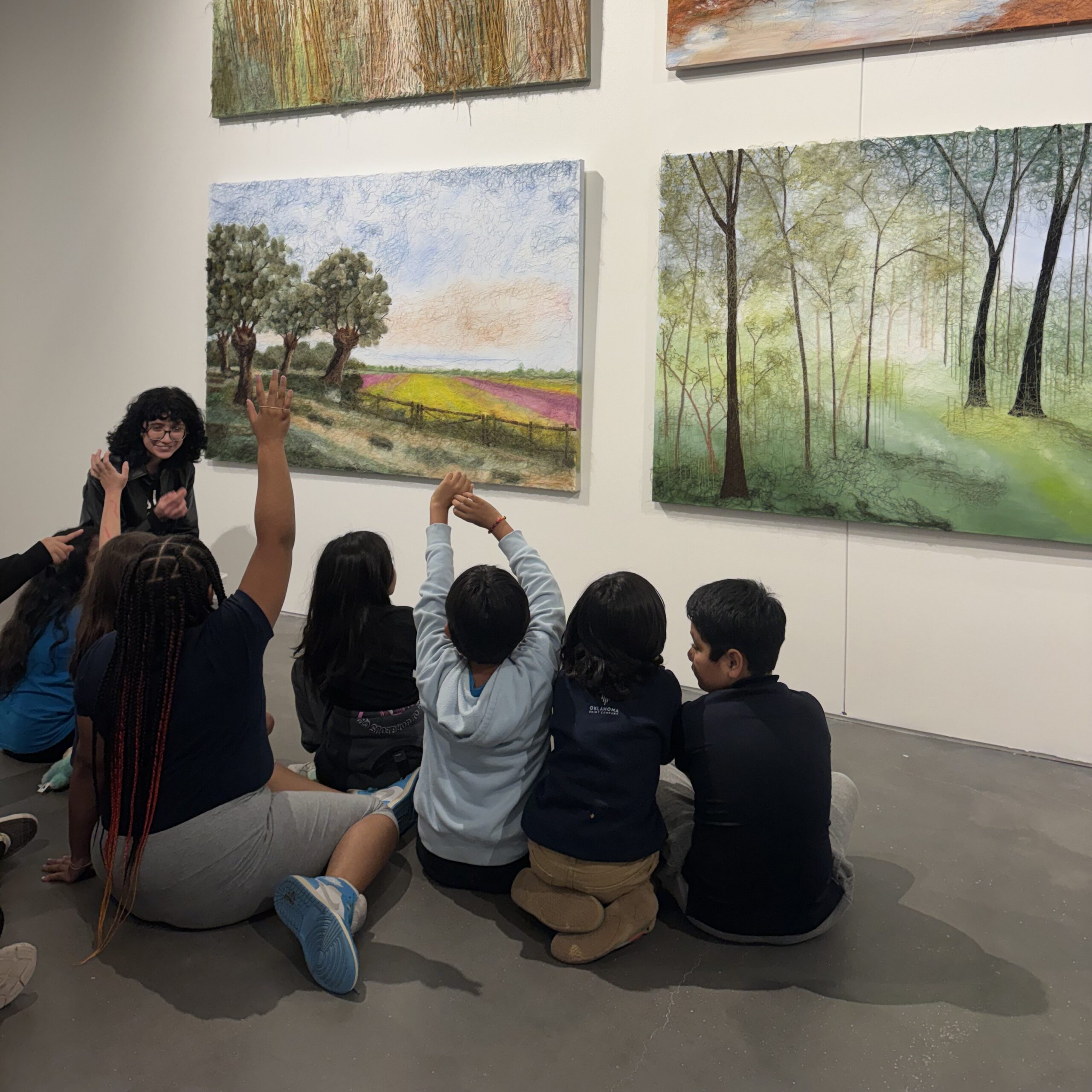 A group of kids sit in front of landscapes with a gallery guide