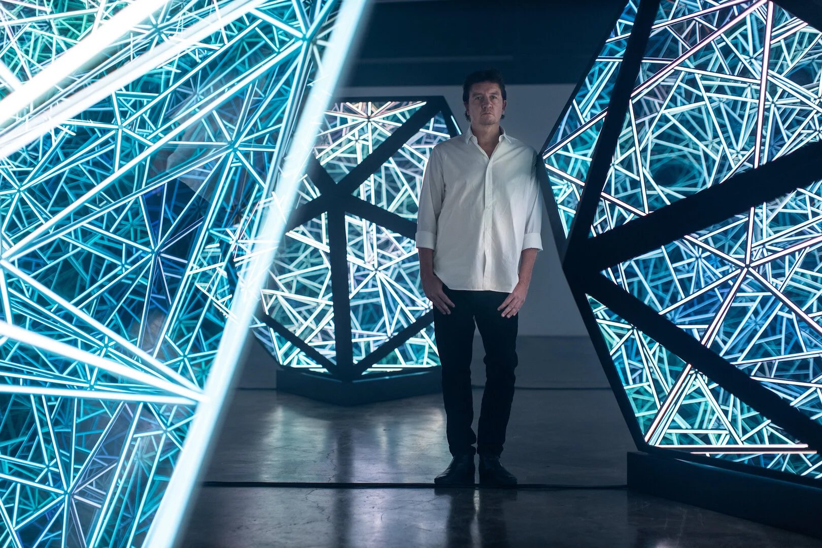 Anthony James, a man in a white shirt and black pants, stands among illuminated, geometric sculptures